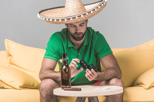 Homem criminoso no sombrero mexicano no sofá amarelo com arma, charuto e garrafa de cerveja em cinza — Fotografia de Stock