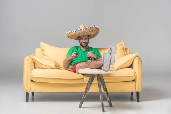 Happy man in mexican sombrero with bottle of beer watching TV on yellow sofa on grey — Stock Photo