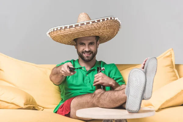 Guapo hombre sonriente en sombrero mexicano viendo la televisión con control remoto mientras está sentado en un sofá amarillo con botella de cerveza en gris — Stock Photo