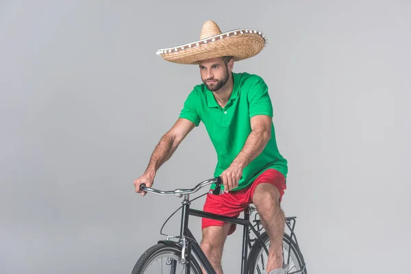 Handsome man in mexican sombrero riding bike on grey — Stock Photo