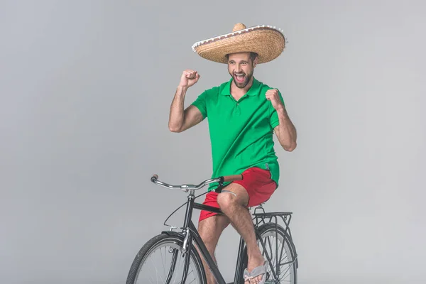 Uomo allegro in sombrero messicano che festeggia e si siede in bicicletta sul grigio — Foto stock