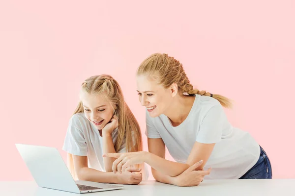 Sonrientes madre e hija en camisetas blancas señalando y mirando a la computadora portátil juntos aislados en rosa - foto de stock