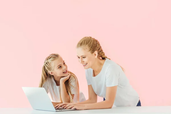 Beautiful mother and daughter in white t-shirts using laptop together isolated on pink — Stock Photo