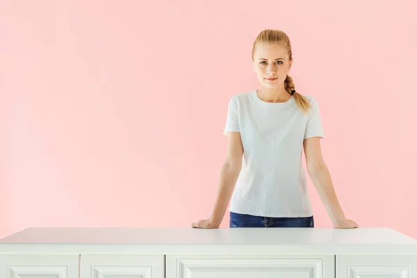 Attraktive junge Frau hinter Küchentisch isoliert auf rosa — Stockfoto