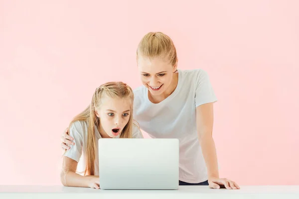 Happy mother and surprised daughter in white t-shirts using laptop together isolated on pink — Stock Photo