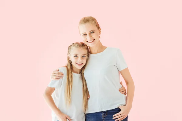 Smiling mother and daughter in white t-shirts embracing and looking at camera isolated on pink — Stock Photo