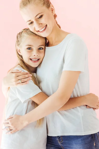 Feliz madre e hija en camisetas blancas abrazando y mirando a la cámara aislada en rosa - foto de stock