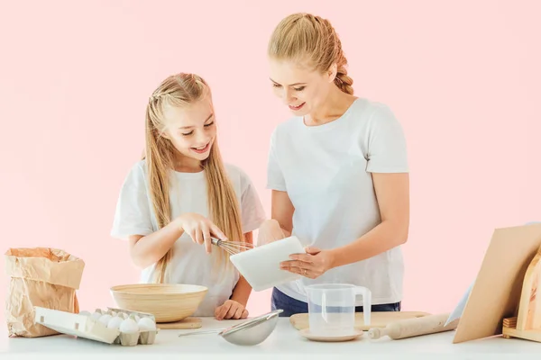 Attractive mother and little daughter in white t-shirts using tablet while cooking isolated on pink — Stock Photo