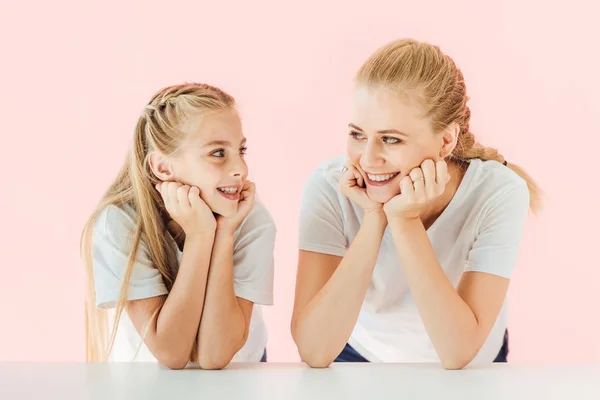 Feliz jovem mãe e filha em camisetas brancas apoiadas na mesa e olhando uns para os outros isolados em rosa — Fotografia de Stock