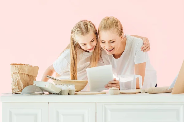 Laughing mother and daughter in white t-shirts using tablet while cooking isolated on pink — Stock Photo