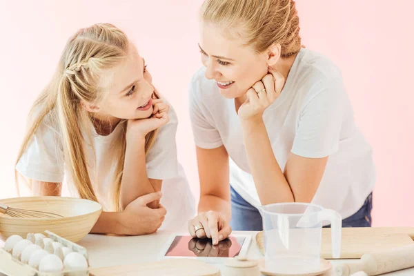 Beautiful mother and daughter in white t-shirts using tablet while cooking together isolated on pink — Stock Photo