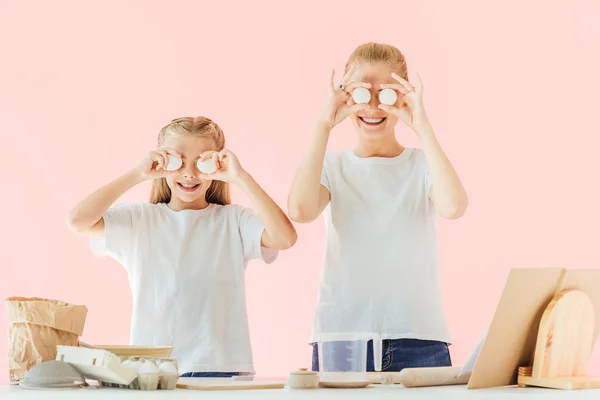 Sonrientes jóvenes madre e hija en camisetas blancas cubriendo los ojos con huevos de pollo mientras cocinan aislados en rosa - foto de stock