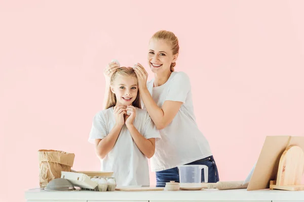 Heureuse jeune mère attachant des œufs de poulet à la tête des filles pour faire des oreilles de lapin tout en cuisinant isolé sur rose — Photo de stock