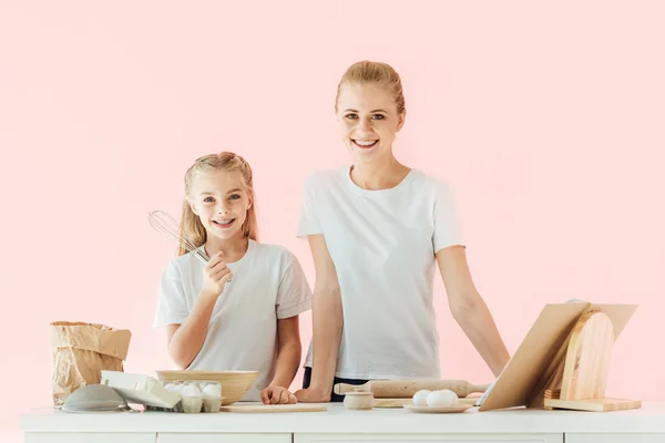 Glückliche Mutter und Tochter in weißen T-Shirts, die beim gemeinsamen Kochen isoliert in rosa in die Kamera schauen — Stockfoto