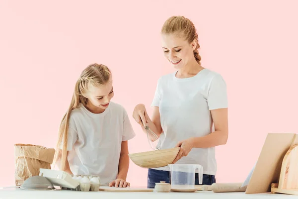 Heureuse mère et fille en t-shirts blancs regardant bol tout en cuisinant ensemble isolé sur rose — Photo de stock