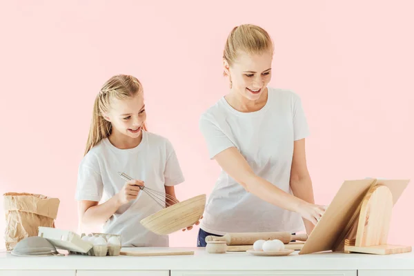 Mãe feliz e filha em camisetas brancas olhando para livro de receitas enquanto cozinhando juntos isolados em rosa — Fotografia de Stock