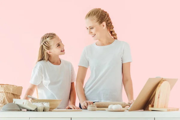 Lächelnde Mutter und Tochter in weißen T-Shirts schauen einander beim gemeinsamen Kochen isoliert auf rosa — Stockfoto