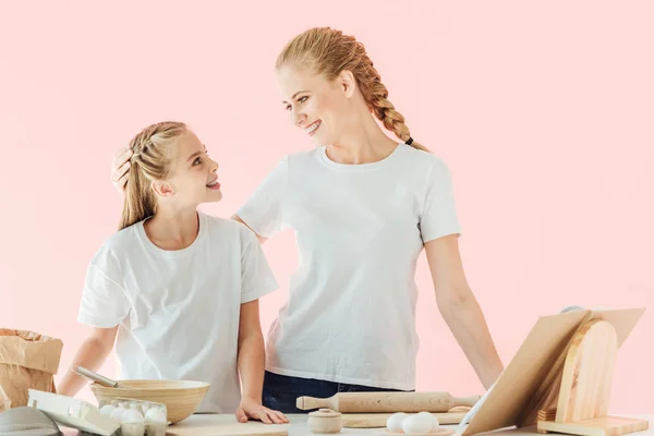 Mãe feliz e filha em camisetas brancas olhando umas para as outras enquanto cozinhavam juntas isoladas em rosa — Fotografia de Stock