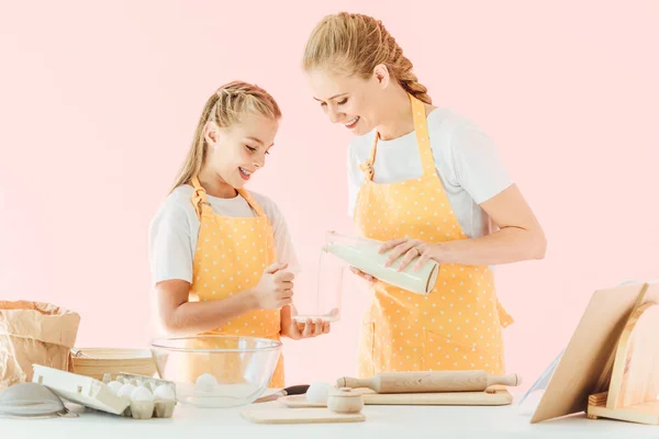 Young mother pouring milk for preparing dough with daughter isolated on pink — Stock Photo