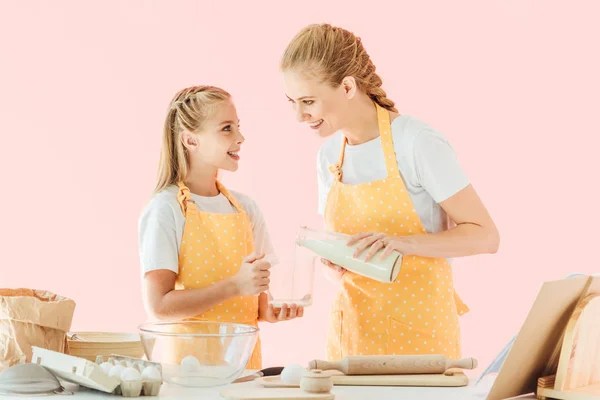 Glückliche Mutter gießt Milch für Teigzubereitung mit Tochter isoliert auf rosa — Stockfoto