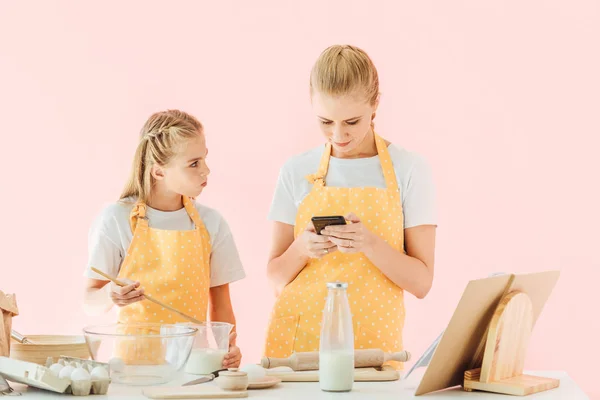 Attractive young mother and daughter using smartphone while cooking together isolated on pink — Stock Photo