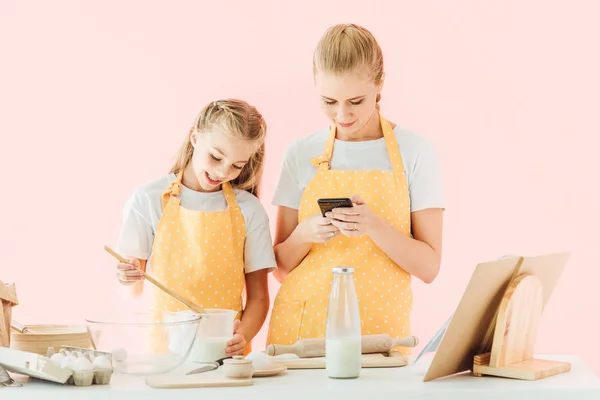 Sonrientes madre e hija usando smartphone mientras cocinan juntas aisladas en rosa - foto de stock