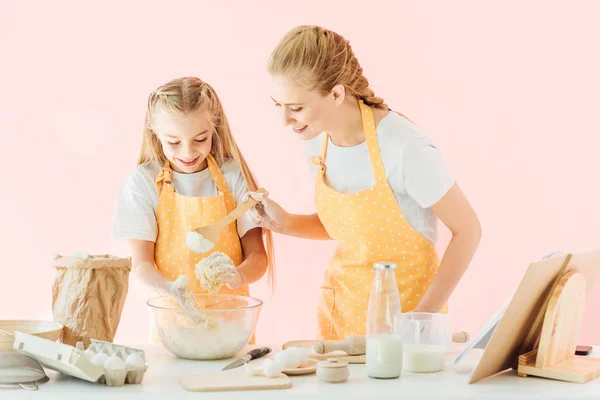Счастливая молодая мать и очаровательная маленькая дочь в желтых фартуках, которые вместе делают тесто, изолированное на розовом — стоковое фото