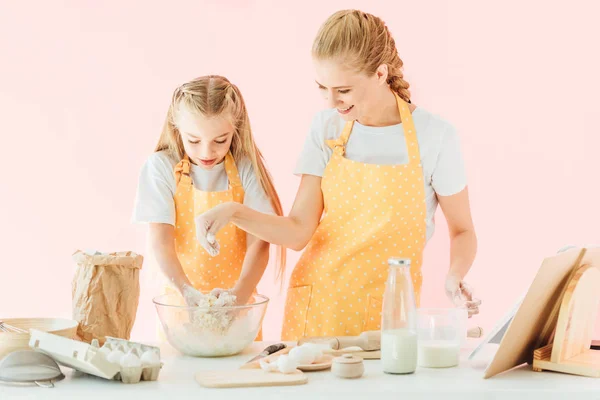 Sonriente joven madre y adorable hijita en delantales amarillos haciendo masa juntos aislados en rosa - foto de stock