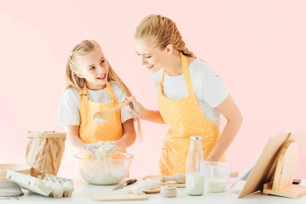 Улыбающаяся молодая мать и очаровательная маленькая дочь в желтых фартуках смешивают тесто, изолированное на розовом — стоковое фото