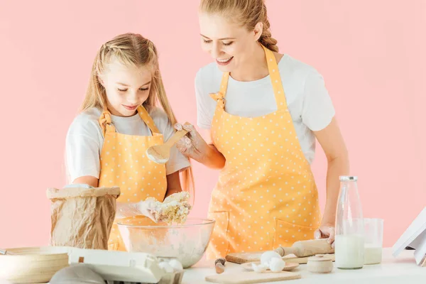 Young mother and adorable little daughter in yellow aprons kneading dough together isolated on pink — Stock Photo