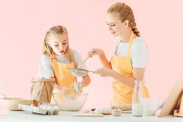 Smiling mother and excited little daughter in yellow aprons preparing dough together isolated on pink — Stock Photo