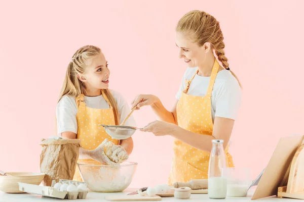 Mãe sorridente e filha em aventais amarelos preparando massa juntos isolados em rosa — Fotografia de Stock