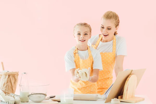 Madre joven sorprendida mirando a la hija amasando masa aislada en rosa - foto de stock