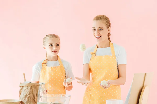 Sonrientes madre e hija haciendo malabares con trozos de masa mientras cocinan aislados en rosa - foto de stock