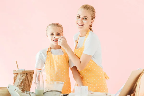 Усміхнена мати і дочка з борошном на обличчях дивиться на камеру під час приготування їжі ізольовані на рожевому — стокове фото
