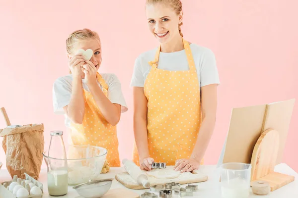 Sonriente joven madre e hija con masa en forma de corazón mirando a la cámara mientras prepara galletas aisladas en rosa - foto de stock