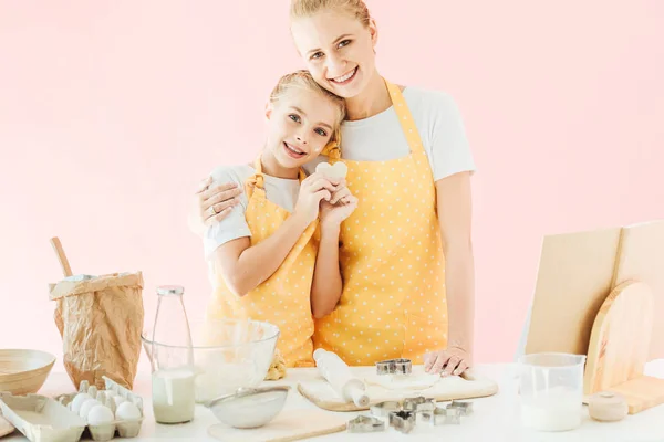 Feliz joven madre e hija con la masa en forma de corazón mirando a la cámara mientras se preparan galletas aisladas en rosa - foto de stock