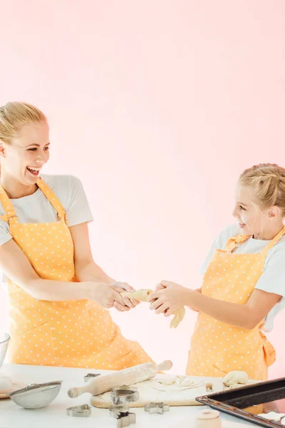 Happy mother and daughter pulling piece of dough together and laughing isolated on pink — Stock Photo