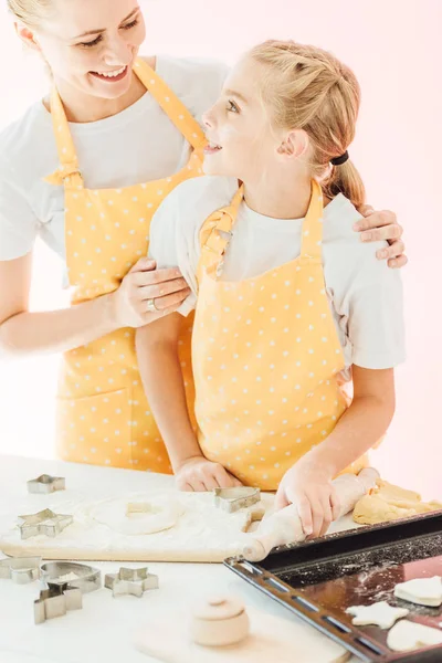 Sorrindo mãe e filha preparando massa para biscoitos juntos e olhando uns para os outros isolados no rosa — Fotografia de Stock