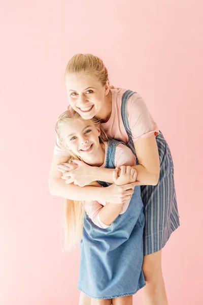 Felice giovane madre e figlia in abiti blu abbracciando e guardando la fotocamera isolata su rosa — Foto stock