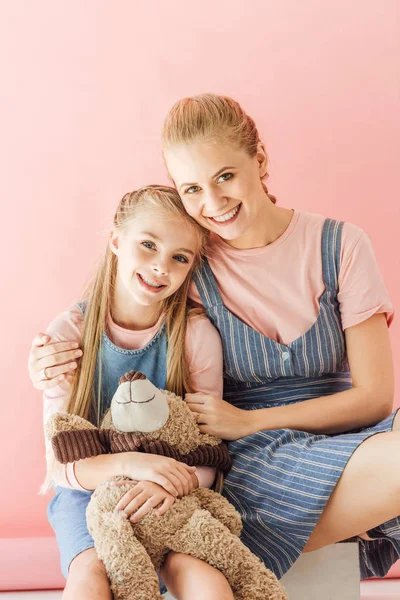 Hermosa feliz madre e hija con osito de peluche abrazando y mirando a la cámara aislada en rosa - foto de stock