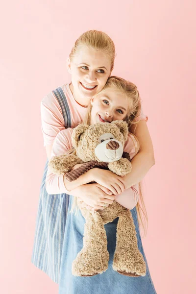 Smiling mother and daughter with teddy bear embracing isolated on pink — Stock Photo