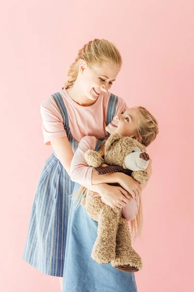 Happy mother and daughter with teddy bear embracing and looking at each other isolated on pink — Stock Photo