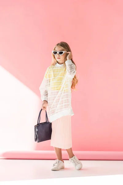 Rubia joven de moda en gafas de sol posando con bolso en rosa - foto de stock