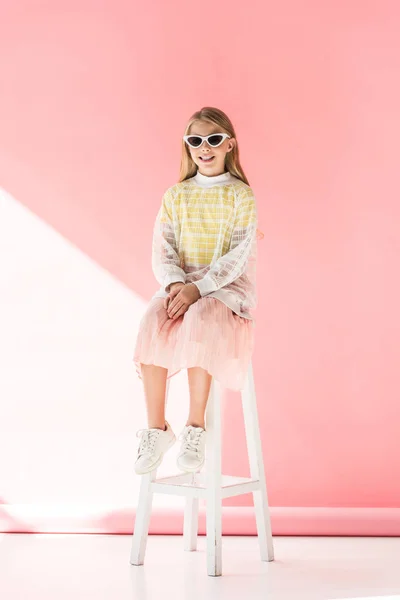 Adorable stylish cheerful kid in sunglasses sitting on stool on pink — Stock Photo