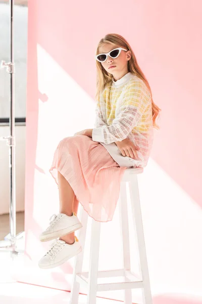 Stylish preteen child in sunglasses sitting on stool on pink — Stock Photo
