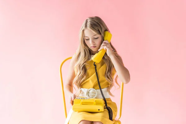 Stylish blonde preteen child talking on retro telephone while sitting on yellow chair on pink — Stock Photo