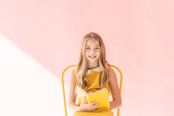 Усміхнена дитина читає книгу, сидячи на жовтому стільці на рожевому — стокове фото