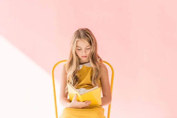 Красива блондинка-молодший читає книгу, сидячи на жовтому стільці на рожевому — стокове фото