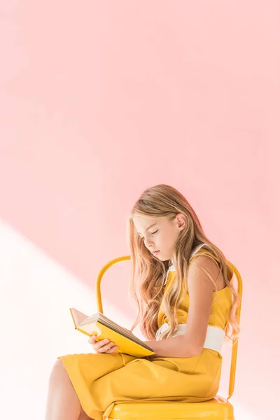 Élégant livre blond enfant de lecture tout en étant assis sur chaise jaune sur rose — Photo de stock